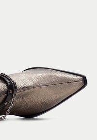Zapato metálico de punta afilada con una superficie texturizada, ribete negro y una cadena decorativa alrededor de la correa del tobillo.