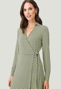 Langearm wikkeldress in een geometrisch patroon met gele, zwarte en witte diamanten. Zacht materiaal met een v-halslijn en strikdetail bij de taille.