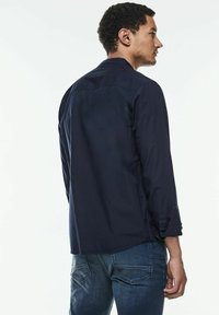 Camisa de manga larga azul marino con puños abotonados, dobladillo recto y detalle en la yugo trasero, combinada con jeans de mezclilla azul oscuro.