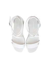 Valkoiset sandaalit, joissa on kaksi nauhaa ja nilkkaremmityyli, jossa on sileä pinta ja pieni koristeellinen metallikoriste sivussa.