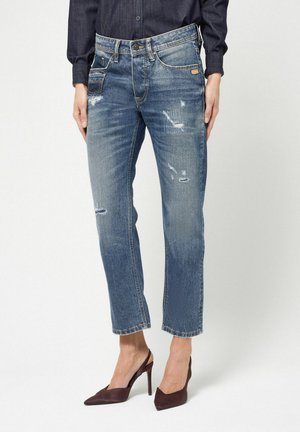 Frau trägt blaue, abgenutzte Straight-Leg-Jeans und dunkle spitze High-Heel-Slingback-Schuhe, steht vor einem weißen Hintergrund.
