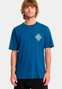 Joven con cabello castaño claro que lleva una camiseta de manga corta azul con un pequeño logo geométrico blanco en el lado izquierdo del pecho, pantalones negros y una pulsera verde.