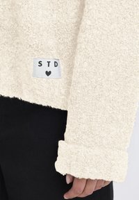 Krém színű bolyhos pulóver, puha, textúrált felülettel és hajtott mandzsettával. Fehér címkéje van, rajta "STD" felirattal és egy szív ikonnal.