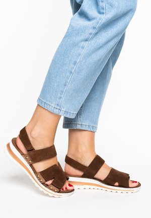 Braune Wildleder-Sandalen mit breiten Riemen, einer gepolsterten Sohle und dekorativen Metallakzenten. Getragen zu hellblauen, verkürzten Jeans.