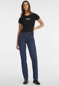 T-shirt noir ajusté avec le logo "GUESS", associé à un jean bleu foncé à jambes droites et des bottes noires, sur un fond blanc uni.