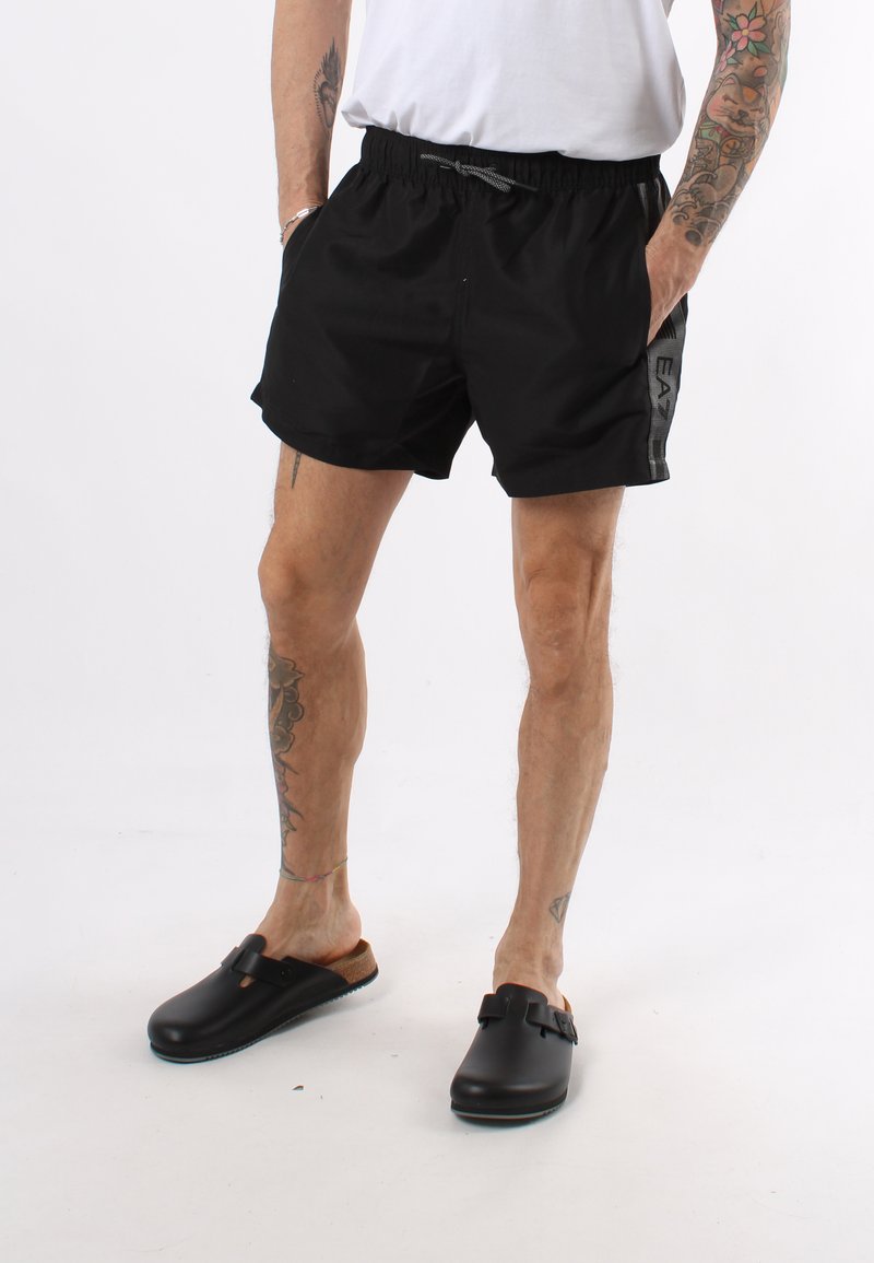 Pantaloncini da bagno neri con elastico in vita e tasche laterali, con marchio grigio. Indossati con zoccoli neri senza lacci.