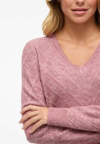 Maglione rosa a scollo a V realizzato in morbido materiale knit, caratterizzato da un motivo a zigzag e polsini a coste. Vestibilità leggermente rilassata con una finitura testurizzata.