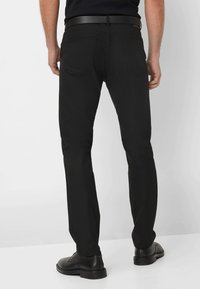 Zwarte broek met een slim fit ontwerp, met een gladde textuur, twee achterzakken en een zichtbaar gestikte tailleband. Draagt met zwarte schoenen.