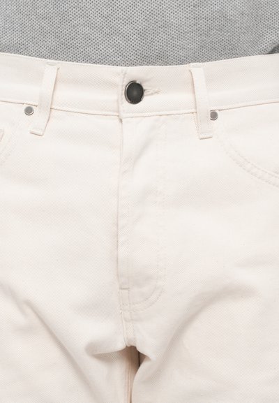 Pantalones de mezclilla de algodón color beige con cierre de cremallera y botón negro, que cuentan con dos bolsillos frontales y una tela suave y texturizada.