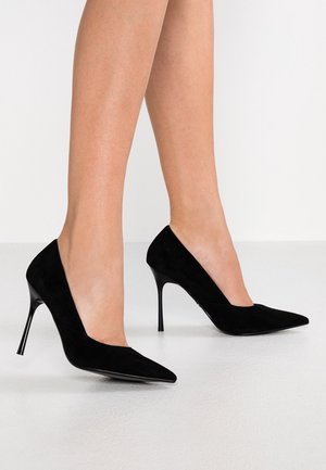 Zwarte suède puntige hoge hakken gedragen op blote voeten, die slanke benen tonen tegen een simpele lichte achtergrond.