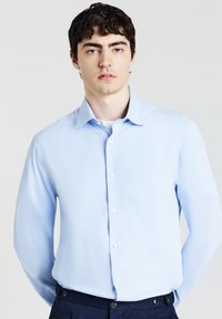 Junger Mann mit dunklen Haaren, der ein hellblaues, geknöpftes Hemd und dunkle Hosen trägt und vor einem schlichten, hellen Hintergrund steht.
