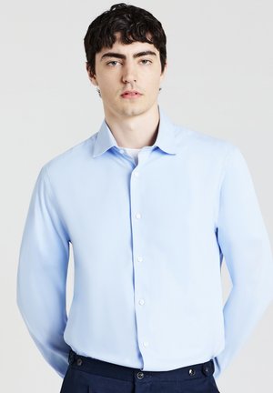 Giovane uomo con capelli scuri, che indossa una camicia azzurra chiara con bottoni e pantaloni scuri, in piedi davanti a uno sfondo chiaro e uniforme.