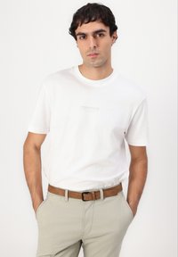 T-shirt bianca in cotone con maniche corte, collezione girocollo, che presenta un piccolo etichetta stampata al centro. Indossata con pantaloni beige chiaro e una cintura marrone.