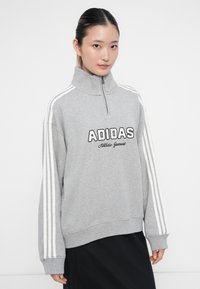 Jovem mulher vestindo um sweatshirt cinza da Adidas com fecho e riscas brancas nas mangas e uma saia preta, de pé contra um fundo branco.