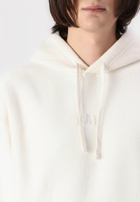Hanorac din fleece de culoare crem, cu o textură moale, dotat cu șnur și un logo brodat subtil. Design cu glugă și croială lejeră.