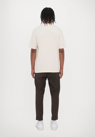 Camiseta de algodón beige de manga corta combinada con pantalones marrón oscuro y zapatillas blancas; puños enrollados visibles en los pantalones.