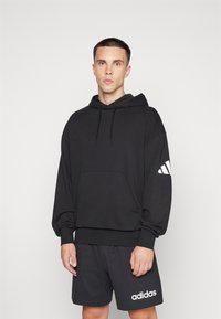 Junger Mann trägt schwarzen Adidas-Hoodie mit weißem Logo am Ärmel und schwarze Adidas-Shorts mit weißem Logo, stehend vor schlichtem hellem Hintergrund.