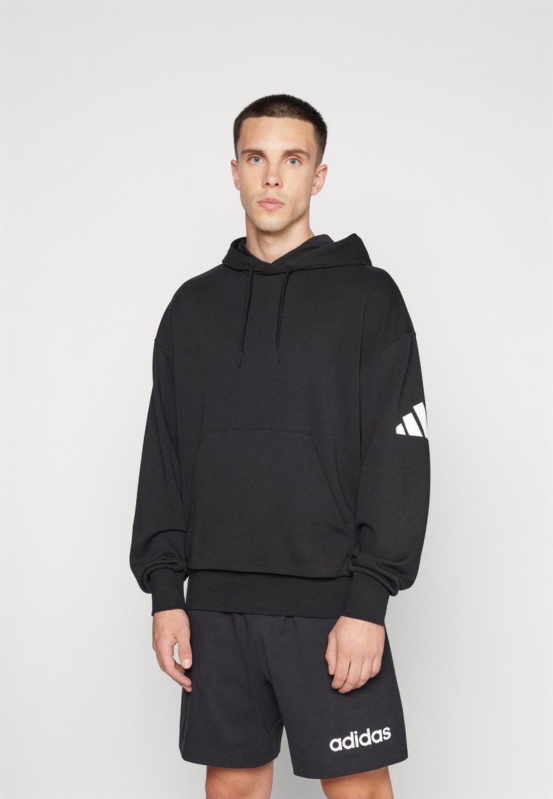 Junger Mann trägt schwarzen Adidas-Hoodie mit weißem Logo am Ärmel und schwarze Adidas-Shorts mit weißem Logo, stehend vor schlichtem hellem Hintergrund.