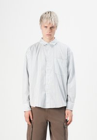 Striped button-up shirt in light blue with red stripes, featuring a pointed collar, single chest pocket, and loose fit, paired with brown pants.