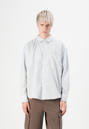 Chemise à rayures bleu clair avec des rayures rouges, dotée d'un col pointu, d'une poche poitrine unique et d'une coupe ample, associée à un pantalon marron.