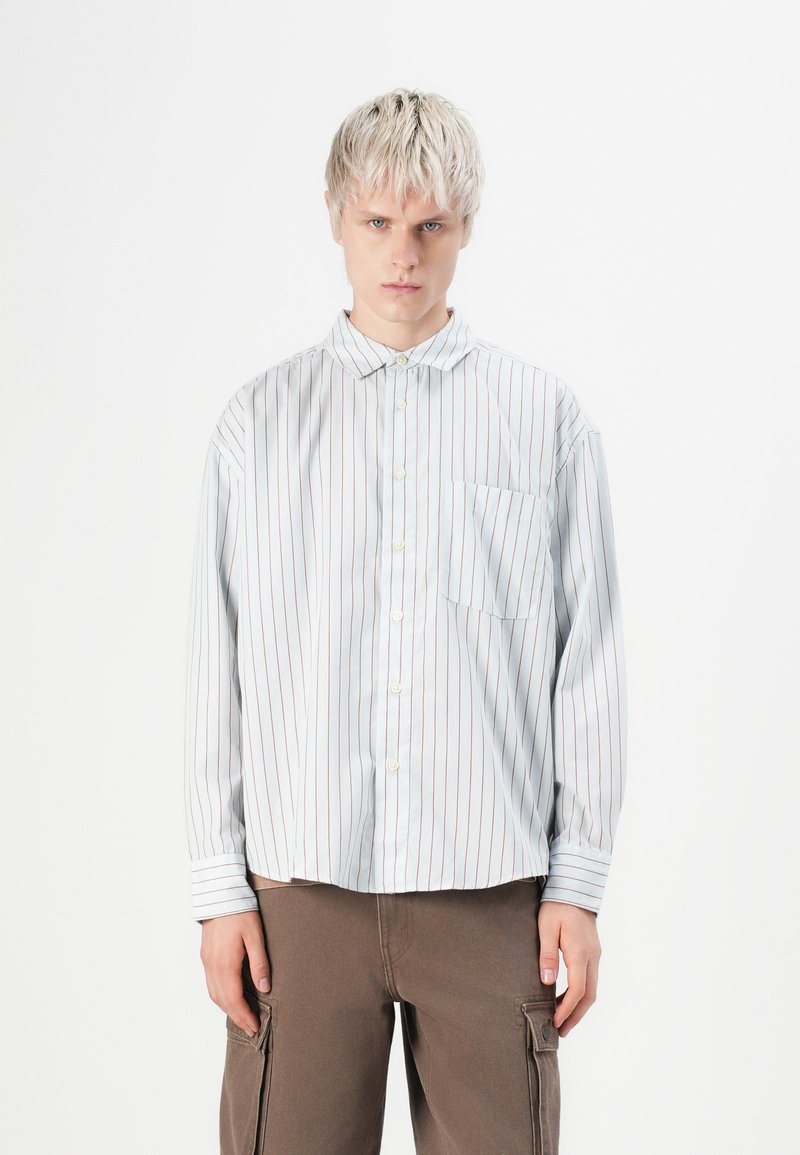 Striped button-up shirt in light blue with red stripes, featuring a pointed collar, single chest pocket, and loose fit, paired with brown pants.