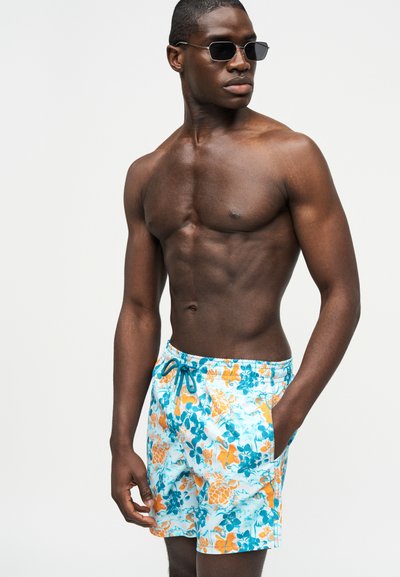 Άνδρας χωρίς πουκάμισο, φοράει μαύρα γυαλιά ηλίου και μπλε μαγιό με πορτοκαλί λουλούδια, με το χέρι στην τσέπη, κοιτάζοντας προς το πλάι.