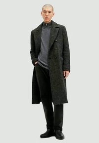 Man wearing a dark gray double-breasted long coat, gray zip-neck sweater, black pants, and black boots, standing with one hand in pocket.