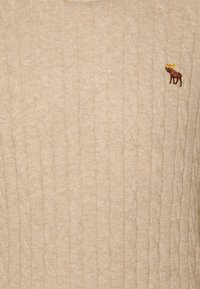 Béžový svetr s pleteným vzorem a malým vyšívaným hnědým losím motivem se žlutými parohy na pravé horní straně hrudi.