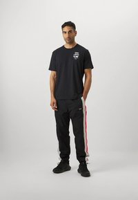 Černé bavlněné tričko s bílým logem na hrudi, doplněné černými tepláky s červeno-šedými bočními pruhy. Kombinováno s černými teniskami.