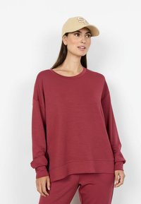 Hanorac supradimensionat de culoare burgundy, cu mâneci lungi și decolteu rotund; asortat cu pantaloni la fel. Șapcă bej cu logo brodat.