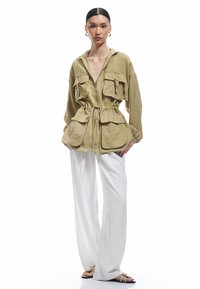 Zielono-oliwkowa kurtka na zamek z przednimi kieszeniami, wcięciem w talii i długimi rękawami, w połączeniu z białymi spodniami o szerokich nogawkach i sandałami.