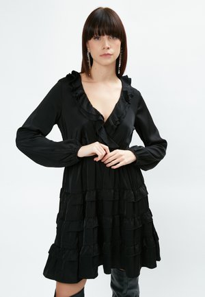 Robe noire à volants avec un col à volants et des manches longues, fabriquée en tissu doux avec un design fluide et une texture plissée.