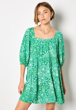 Młoda kobieta ubrana w zieloną i białą kwiecistą krótką sukienkę z bufkami i kwadratowym dekoltem, pozująca na białym tle.
