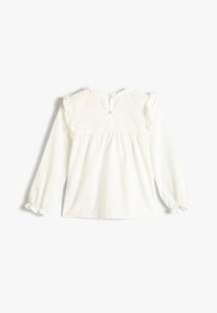 Blouse blanche à manches longues avec des volants au niveau du col et des poignets, comportant une fermeture par bouton au dos et un design ample et froncé.
