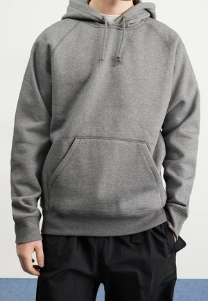 Person iført en ensfarvet grå hættetrøje med forlommer og sorte bukser, hænder delvist synlige ved siderne.