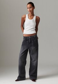 Tmavě šedé džínsy s širokými nohavicemi a stahovacím páskem v pase, doplněné přiléhavým bílým tílkem. Modelka stojí sebevědomě s rukama v bocích.