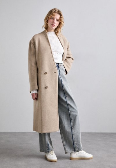 Beige uldfrakke med dobbeltknapper, lange ærmer og afslappet pasform, parret med lyseblå brede jeans og hvide sneakers.