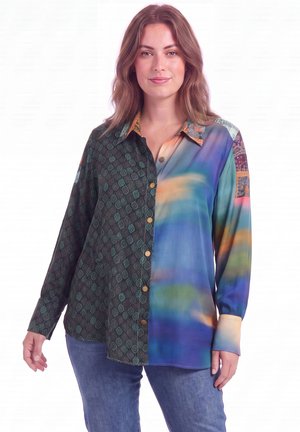 Femme portant une chemise à manches longues boutonnée, avec un côté gauche à motif vert et un motif coloré flou sur le côté droit, associée à un jean bleu.