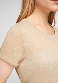 Beige t-shirt med kort ärm gjord av lätt och texturerad tyg med rundad halsringning och uppvikta ärmslut, med subtila färgvariationer.