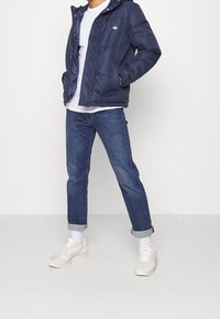 Marinblå puffjacka med huva, dragkedja och quiltad design. Matchas med uppvikta mörka jeans och ljusfärgade sneakers.