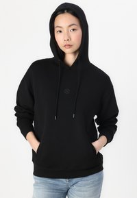 Sweatshirt à capuche noire avec une poche frontale, présentant un petit logo brodé sur la poitrine et des poignets côtelés. Texture de tissu lisse.