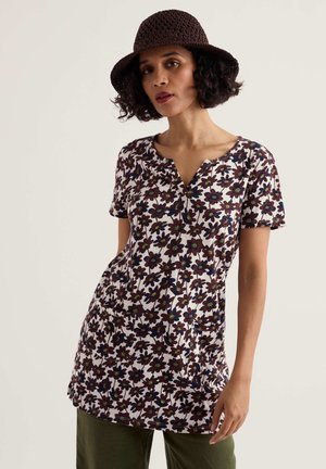 Vrouw draagt een bruin gehaakt hoedje, een tuniek met korte mouwen in een bloemenpatroon en voorzakken, en olijfgroene broek tegen een effen achtergrond.