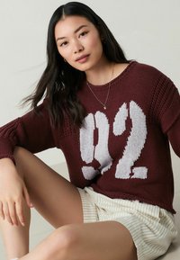 Bordeauxstickad tröja med stort vitt "92"-tryck. Lång passform med texturerade ärmar, parat med ljusrandiga shorts.