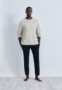 Ανοιχτό γκρι πουλόβερ με ριμπ μανσέτες, σε συνδυασμό με μαύρο παντελόνι με στενή γραμμή. Το σύνολο έχει άνετη εφαρμογή και παρουσιάζεται σε μια όρθια φιγούρα.