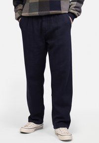 Navy blue relaxed-fit trousers with pleated front, tapered leg, and side pockets, paired with light-colored sneakers.