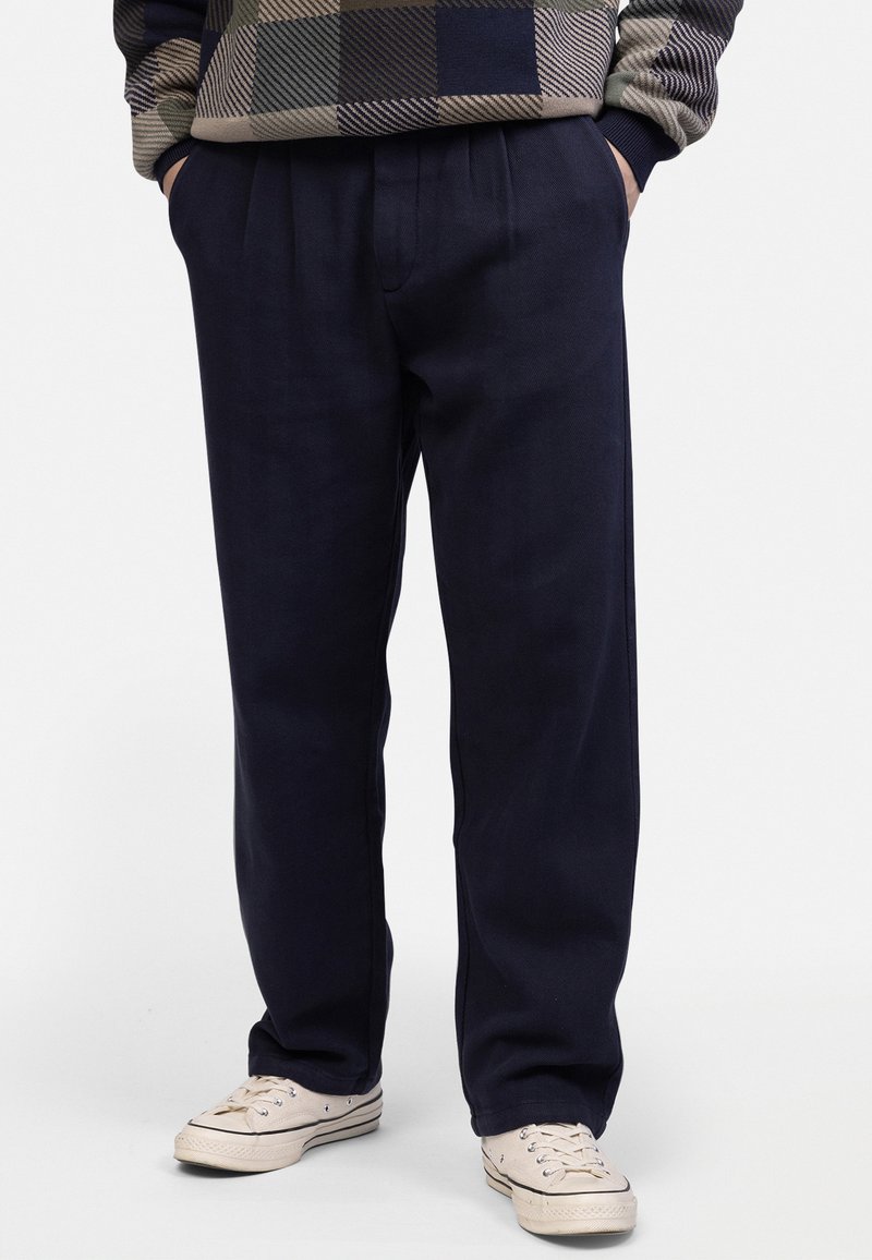 Navy blue relaxed-fit trousers with pleated front, tapered leg, and side pockets, paired with light-colored sneakers.