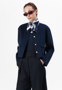 Jachetă din lână cropped de culoare bleumarin cu butoane aurii, două buzunare frontale, asociată cu un top bleumarin și pantaloni negri cu talie înaltă, accesorizată cu o eșarfă cu model.