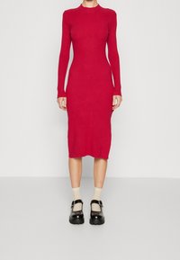 Robe en tricot côtelé rouge avec un col montant et des manches longues, ajustée au corps, longueur sous le genou, associée à des chaussures et des chaussettes noires.