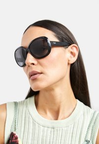 Ochelari de soare negri, supradimensionați, cu lentile întunecate și brațe negre lucioase. Prezintă logo-ul Ray-Ban pe lateral. Modelul poartă un top verde deschis cu nervuri.