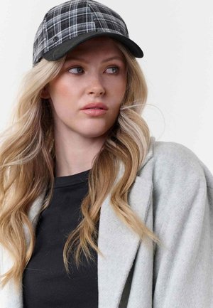 Young woman with long blonde hair wearing a black shirt, grey coat, and black-and-white plaid cap looking to the side against a white background.
